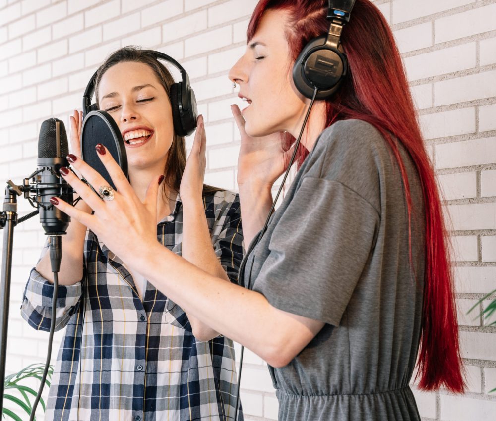 Young women with closed eyes gesticulating and singing into microphone while listening to music in headphones in light professional studio